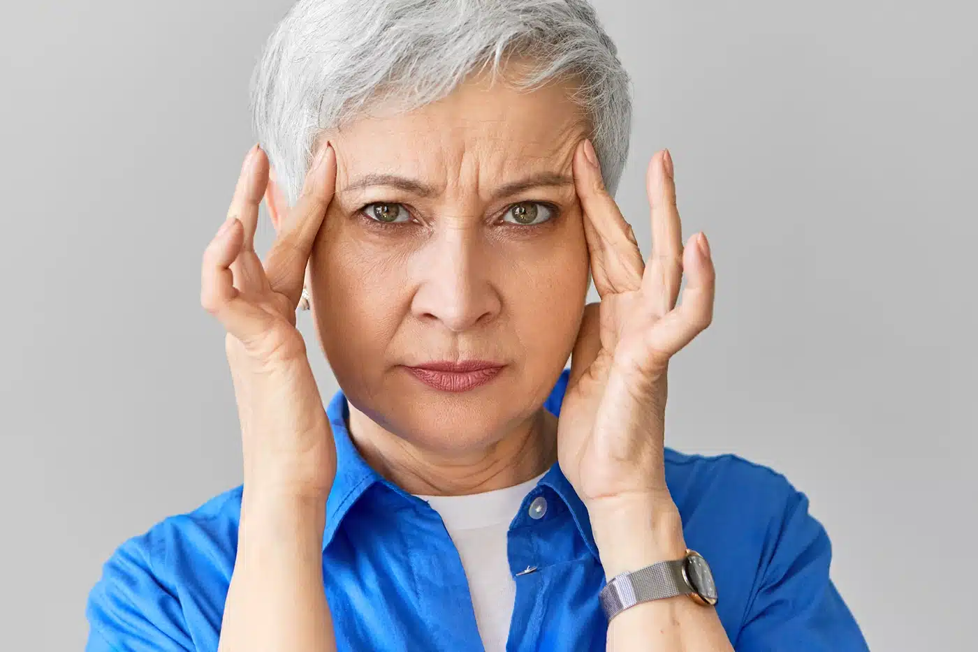 Stylish middle aged Caucasian female in blue shirt suffering from migraine. Close up shot of stressed mature woman squeezing her temples because of terrible headache, massaging painful points